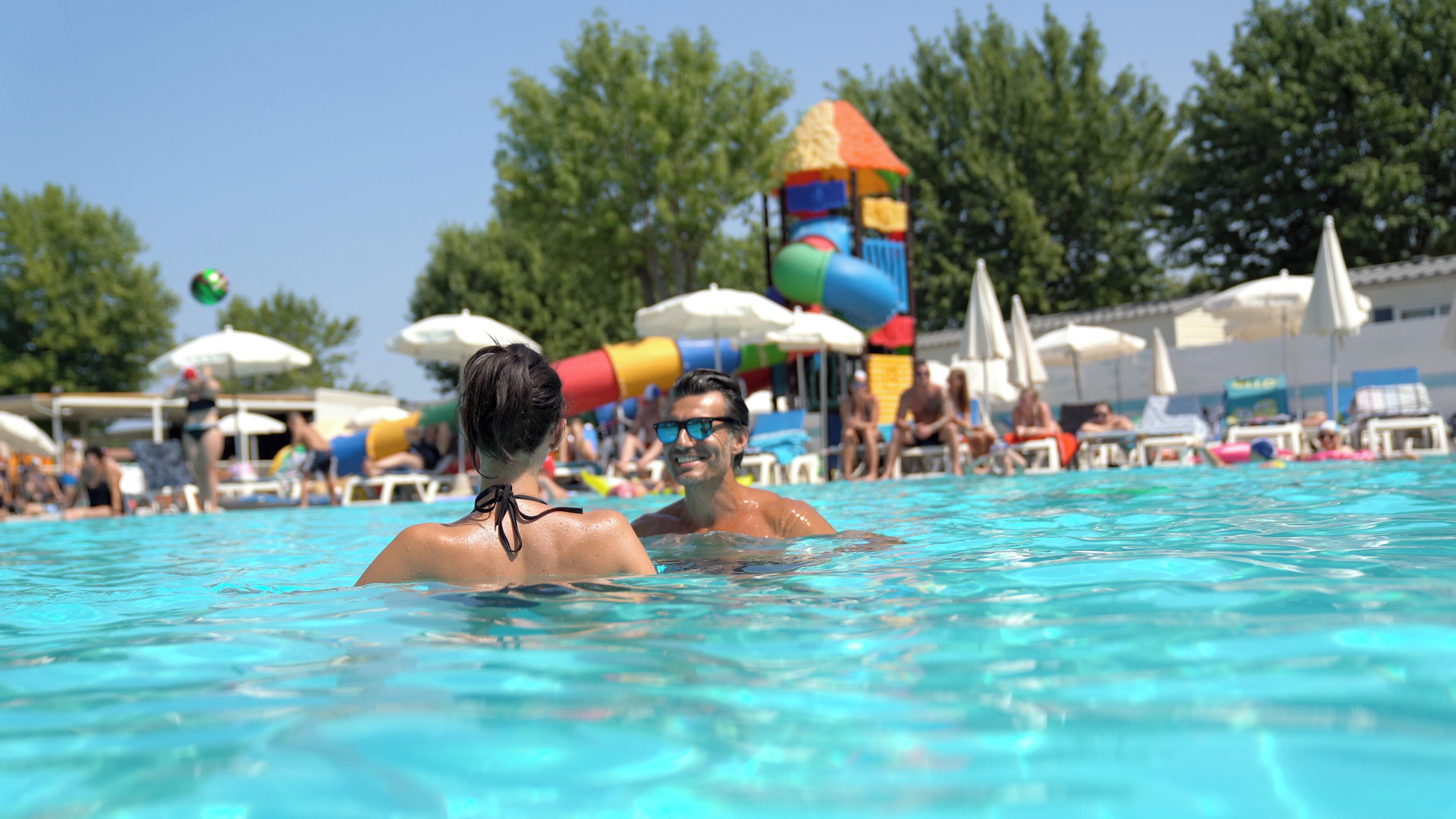 Una piscina con gente en el agua, sombrillas y un tobogán acuático colorido al fondo.