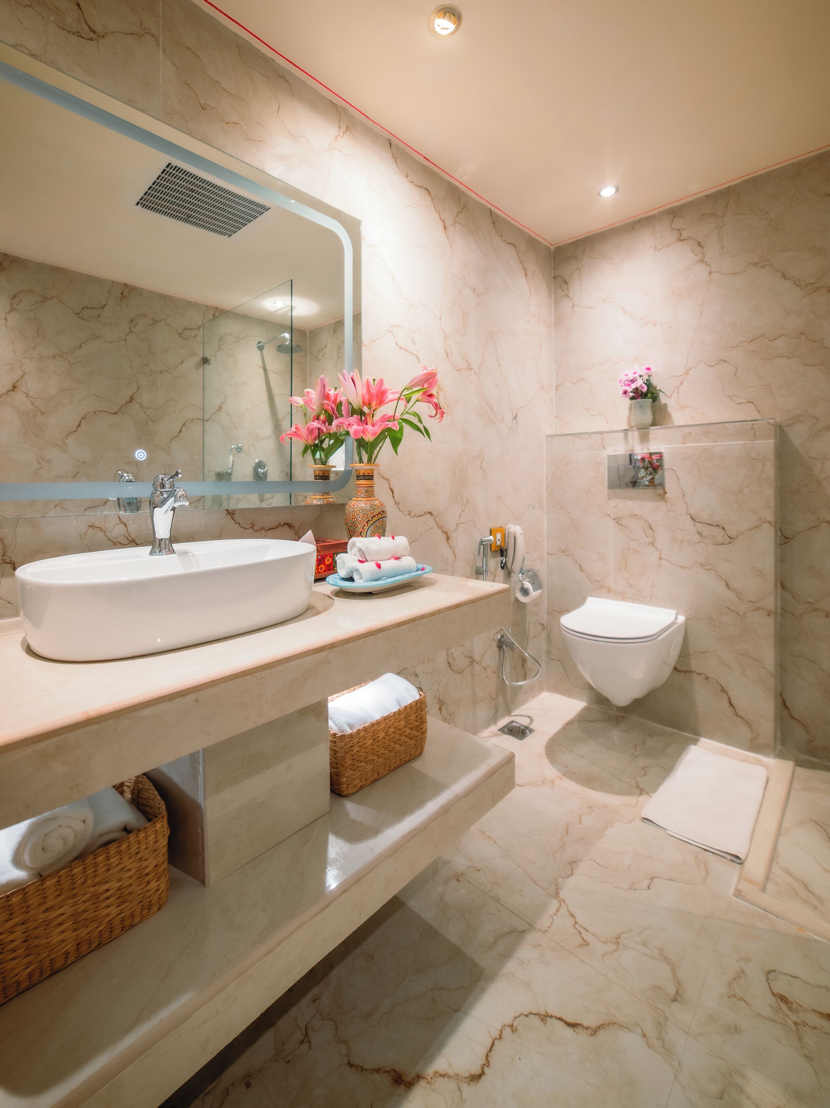 A modern bathroom with a large mirror, a sink, a toilet, and a shower area. There are wicker baskets and a vase with flowers on the counter.