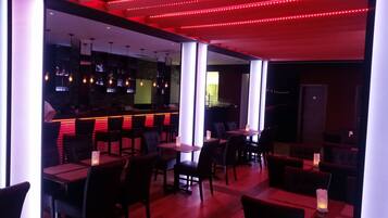 A dimly lit restaurant with red ambient lighting, multiple tables and chairs, and a bar area with a counter and hanging lights.
