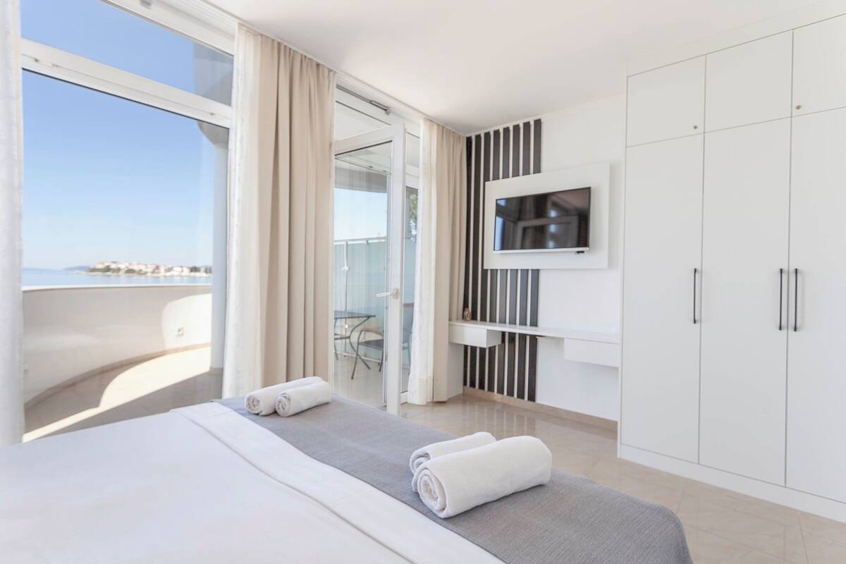 A modern hotel room with a balcony, a bed with rolled towels, a TV mounted on the wall, and a wardrobe.