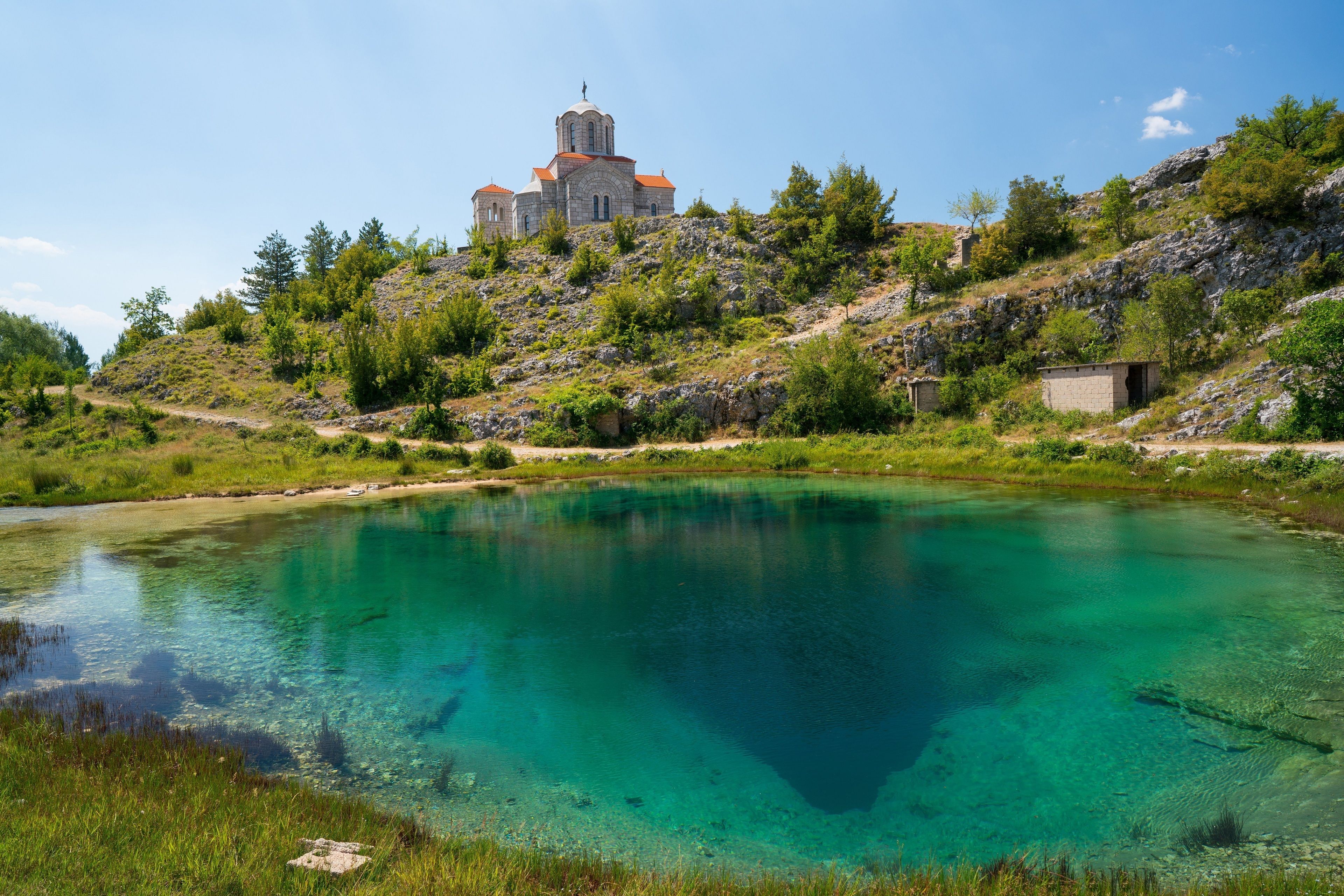 Um lago tranquilo com águas verde-turquesa cristalinas, rodeado por vegetação exuberante e uma colina rochosa com uma igreja.