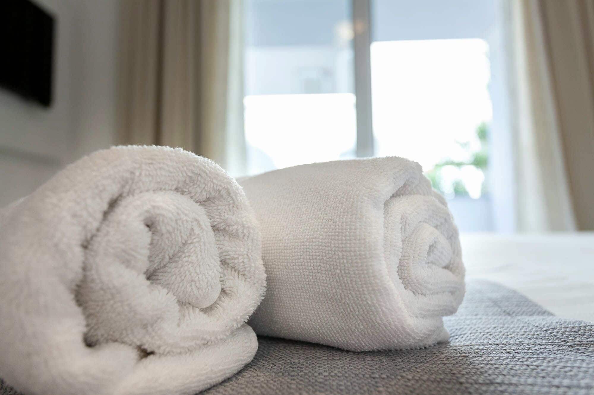 Duas toalhas brancas dobradas sobre uma superfície cinza texturizada, possivelmente uma cama ou sofá, em um quarto com janela e cortinas.