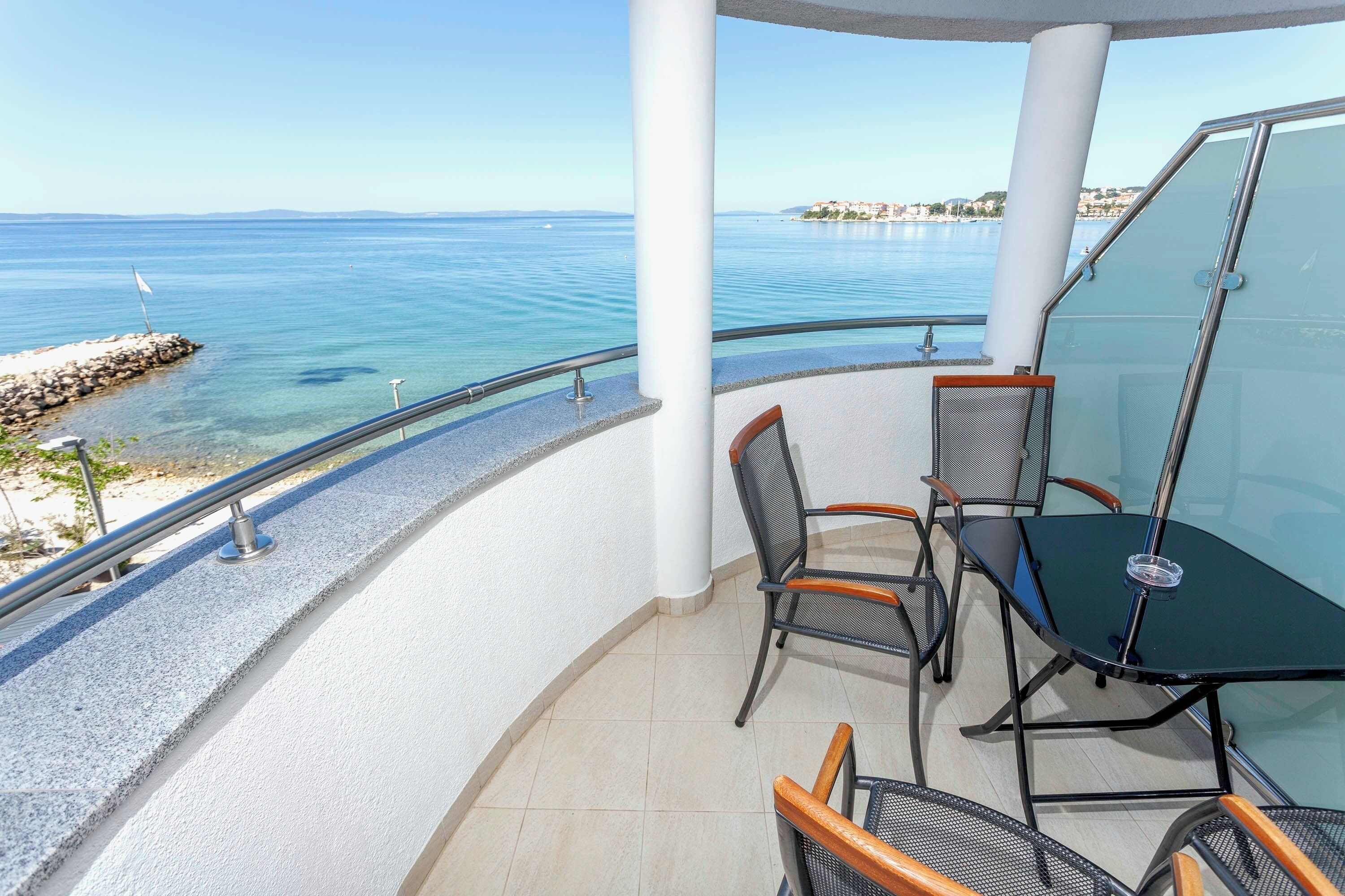 Ein Balkon mit Glastisch und Stühlen, mit Blick auf ein ruhiges Meer und eine entfernte Küstenlinie.