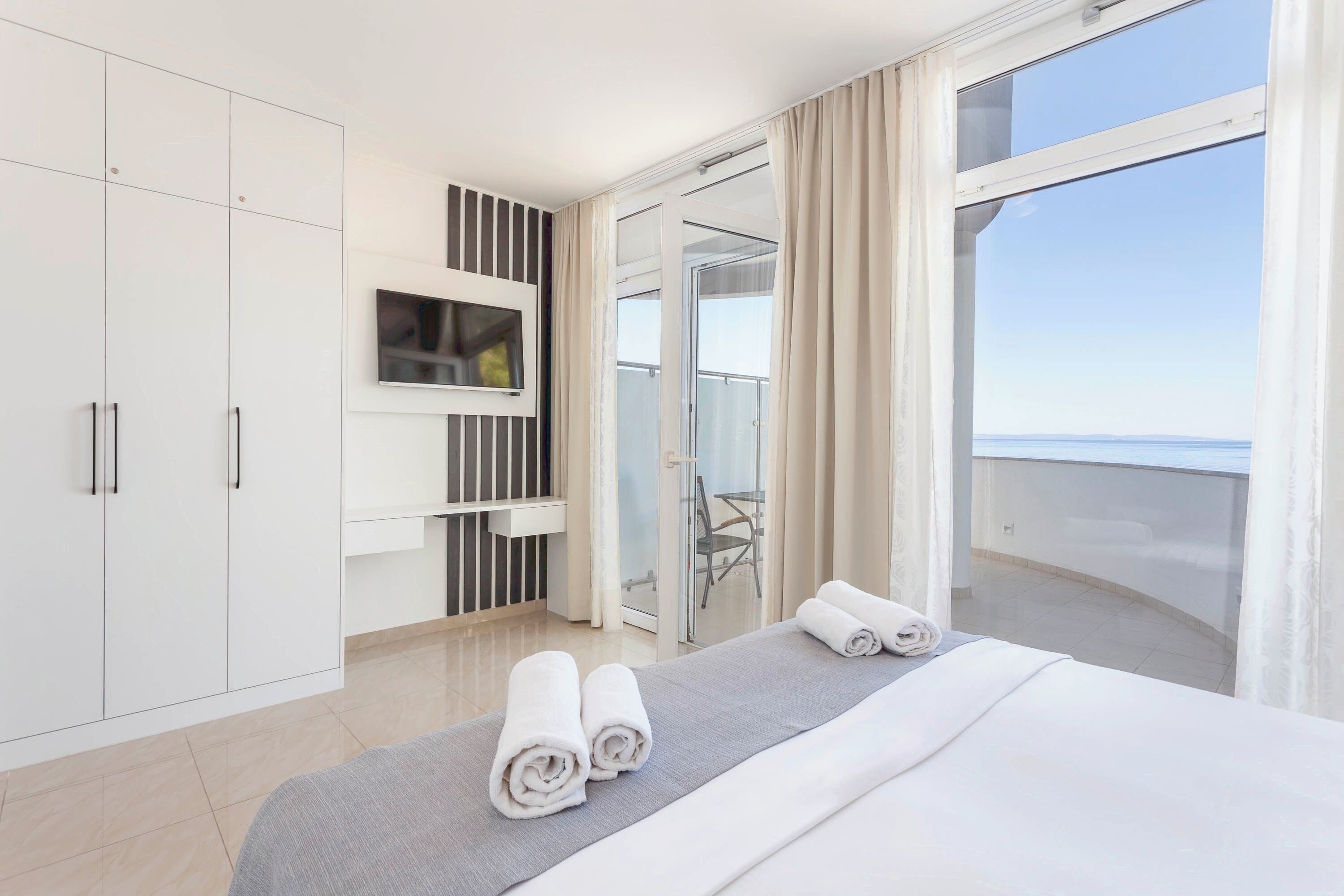Habitación de hotel con cama, televisor montado en la pared, balcón con puerta de cristal y vista al mar.