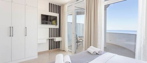 Habitación de hotel con cama, televisor montado en la pared, balcón con puerta de cristal y vista al mar.