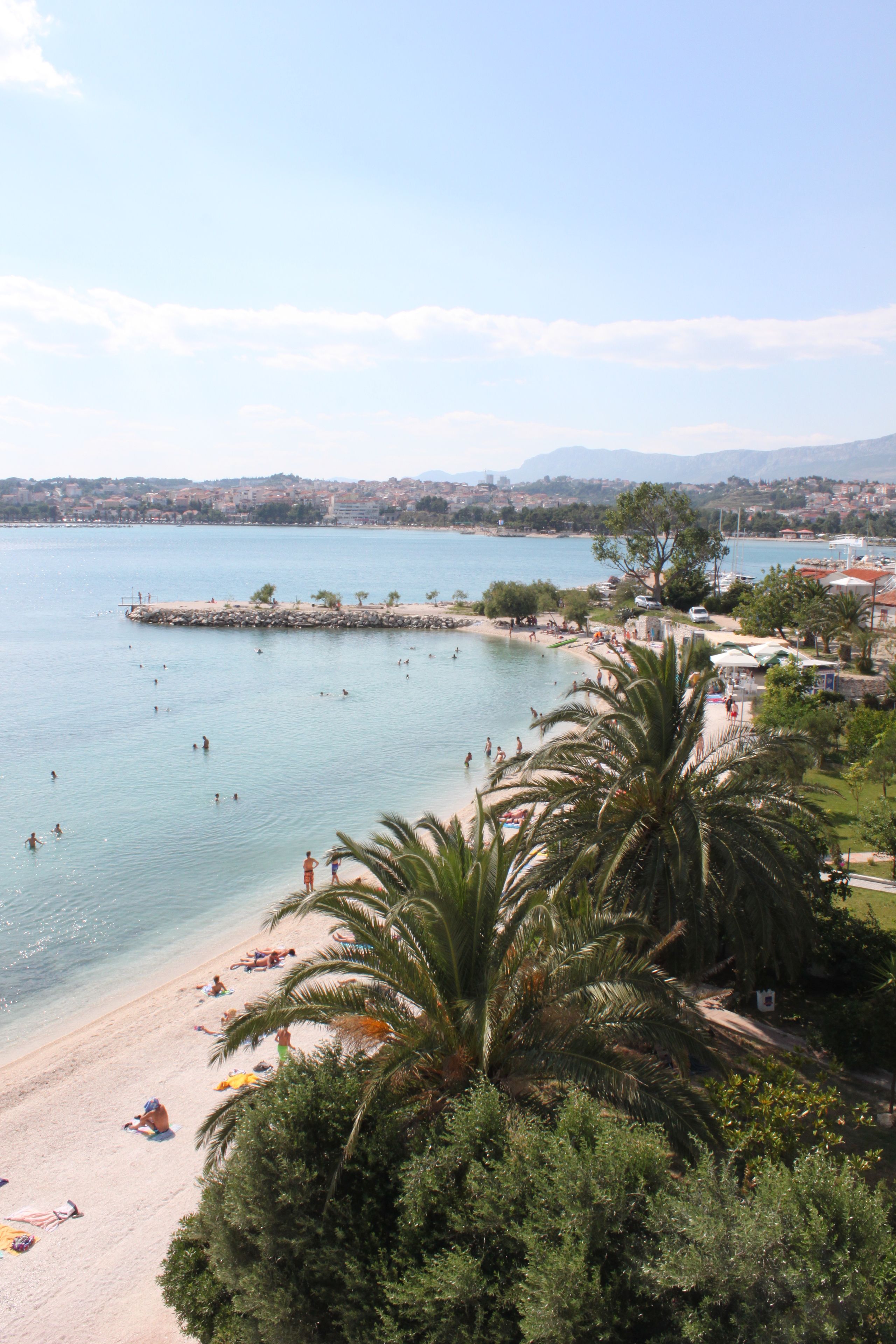 A beach with people swimming and sunbathing, a rocky breakwater, and a cityscape in the background.