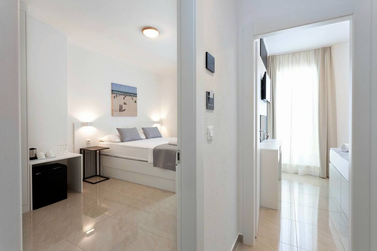 A modern hotel room with a bed, bedside table, and a small desk with a chair. There is a bathroom visible through an open door.