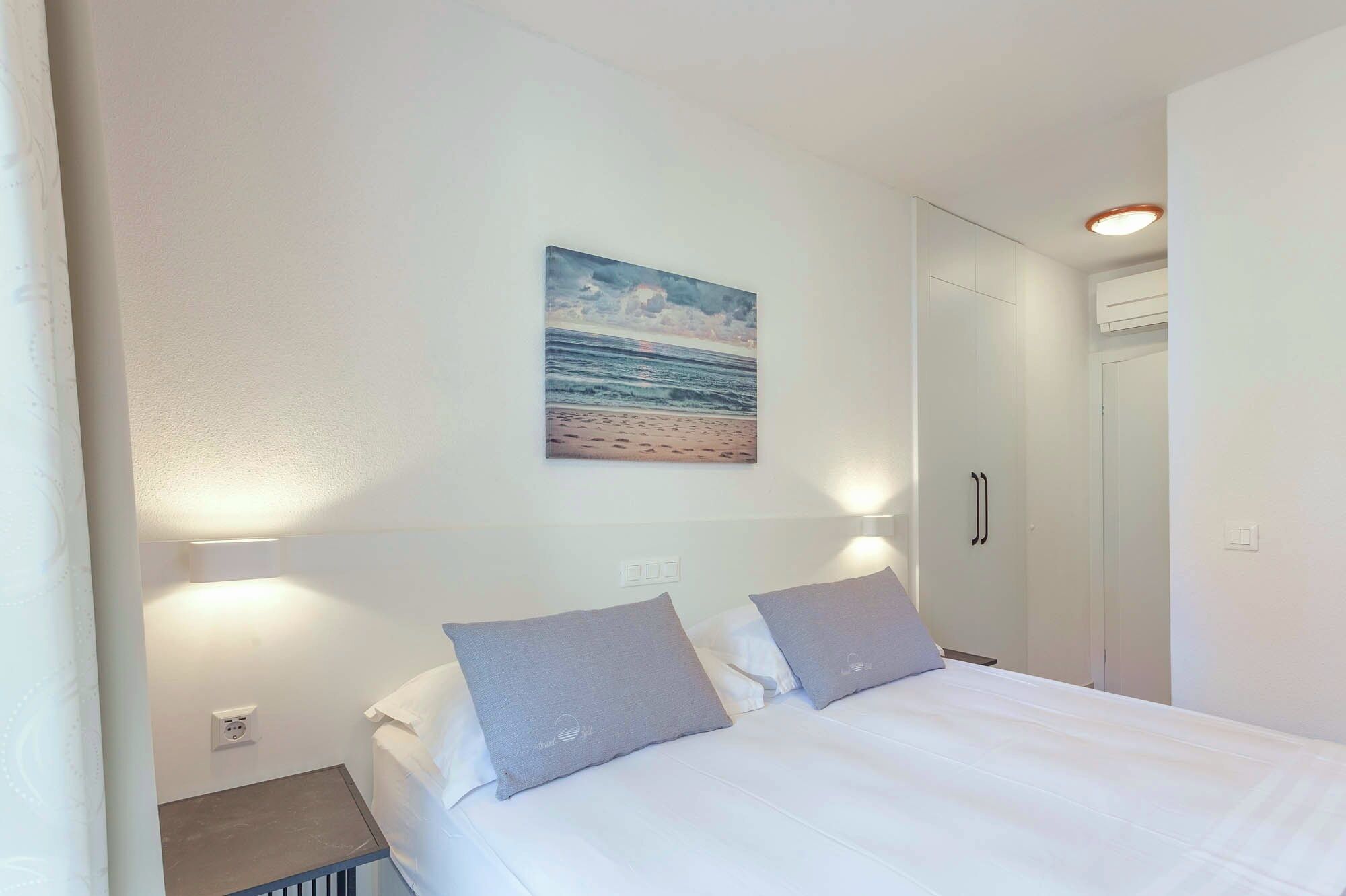 A bedroom with a bed, two pillows, a nightstand, a wall-mounted painting of a beach, and a closet with a door.