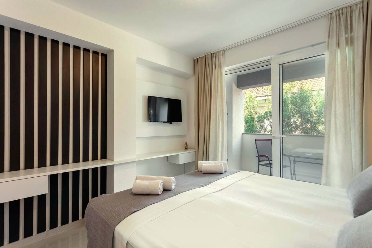 A hotel room with a bed, a TV mounted on the wall, a small table with a chair, and a balcony with curtains.
