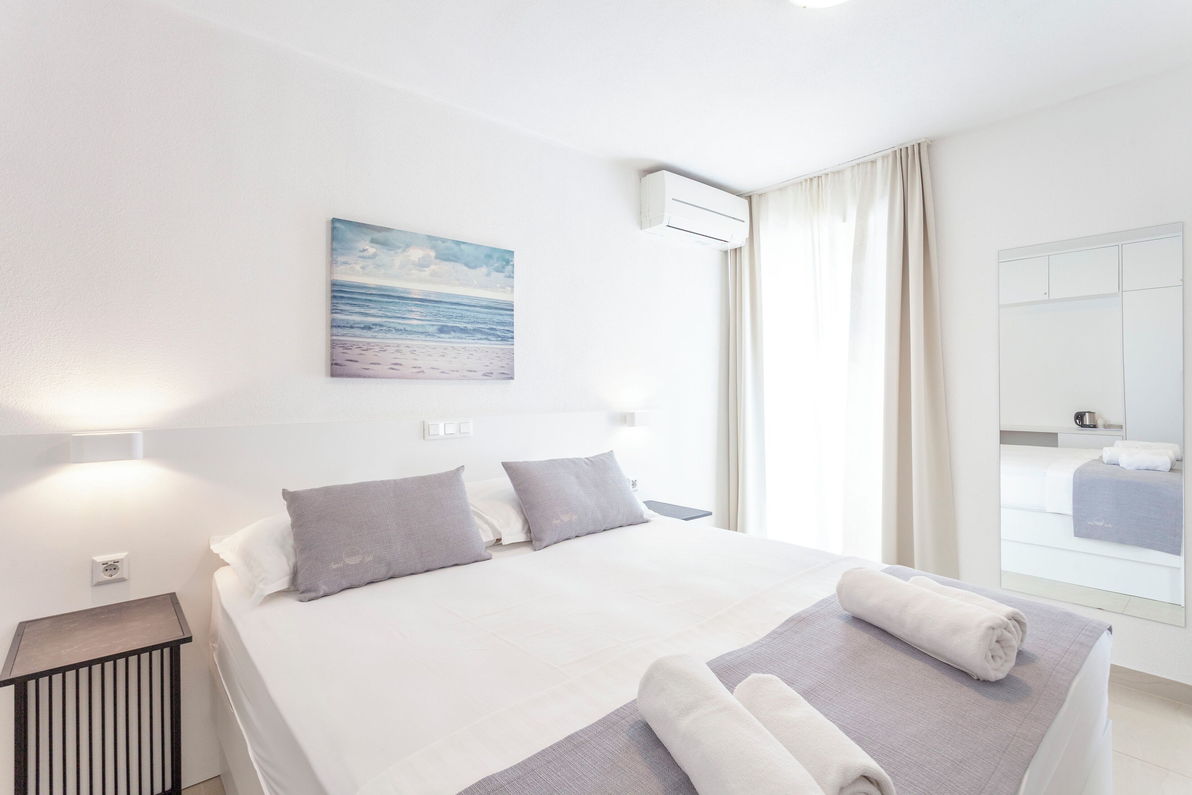 A modern hotel room with a large bed, a painting of a beach, an air conditioning unit, and a view of the room through a window.