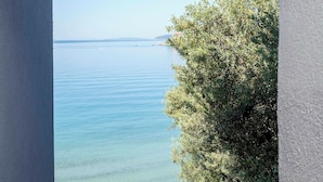 A view through a window of a beach with clear water, a sandy shore, and a tree. - Sunset Split Rooms (Podstrana)