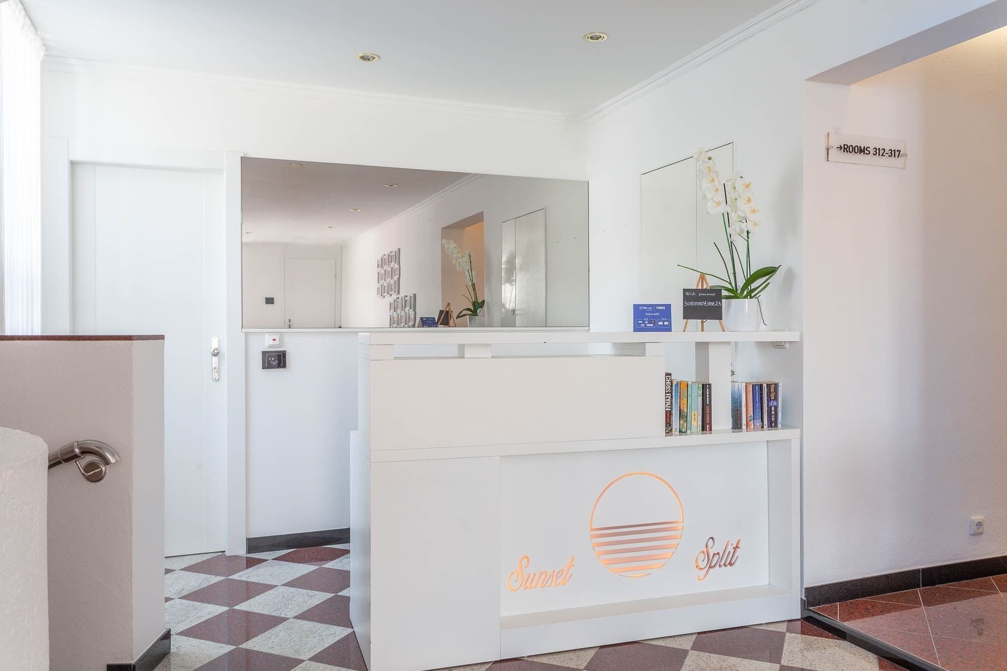 A reception area with a check-in counter, a sign indicating rooms 312-317, a small plant, and a bookshelf with books.