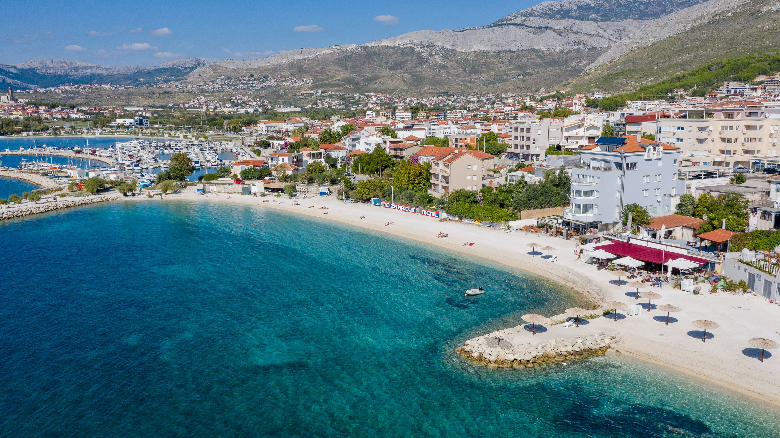 A coastal town with a marina, sandy beach, and buildings with balconies overlooking the sea.