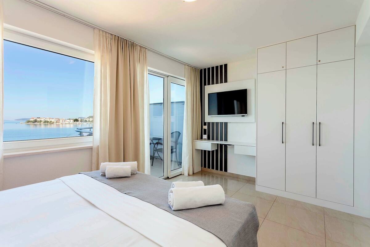 A hotel room with a large bed, a TV mounted on the wall, a wardrobe, and a view of the sea through a large window.