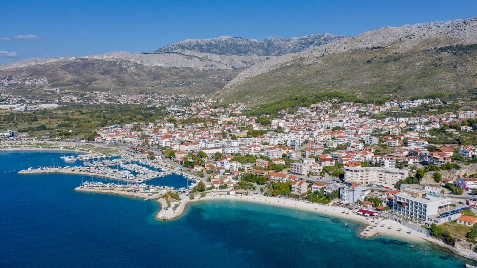 An aerial view of a coastal town with a marina, buildings, and mountains in the background.