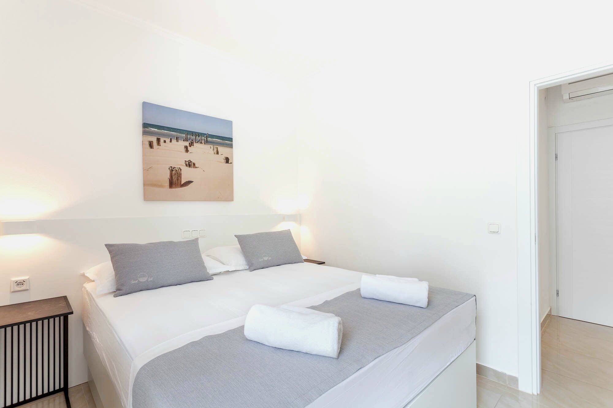 Una habitación de hotel con una cama, una mesita de noche, un cuadro colgado en la pared de una playa y una puerta.