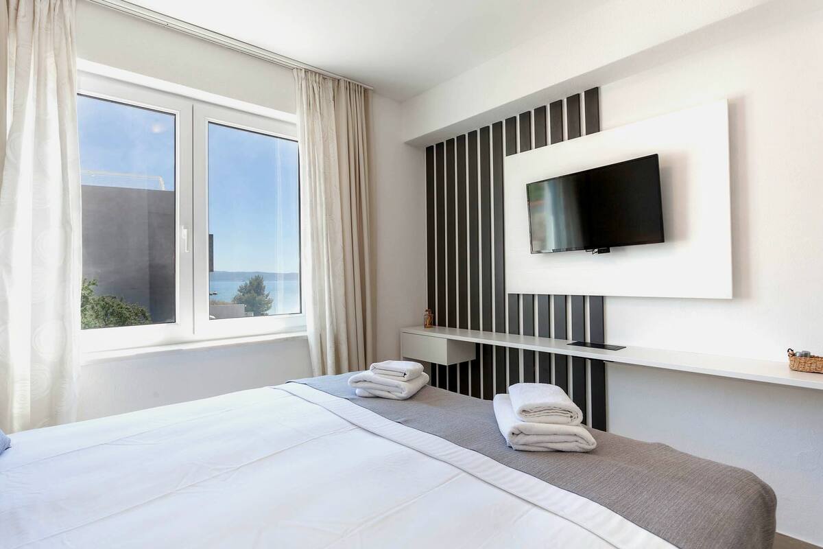 A hotel room with a bed, a TV mounted on the wall, a window with curtains, and a view of the sea.