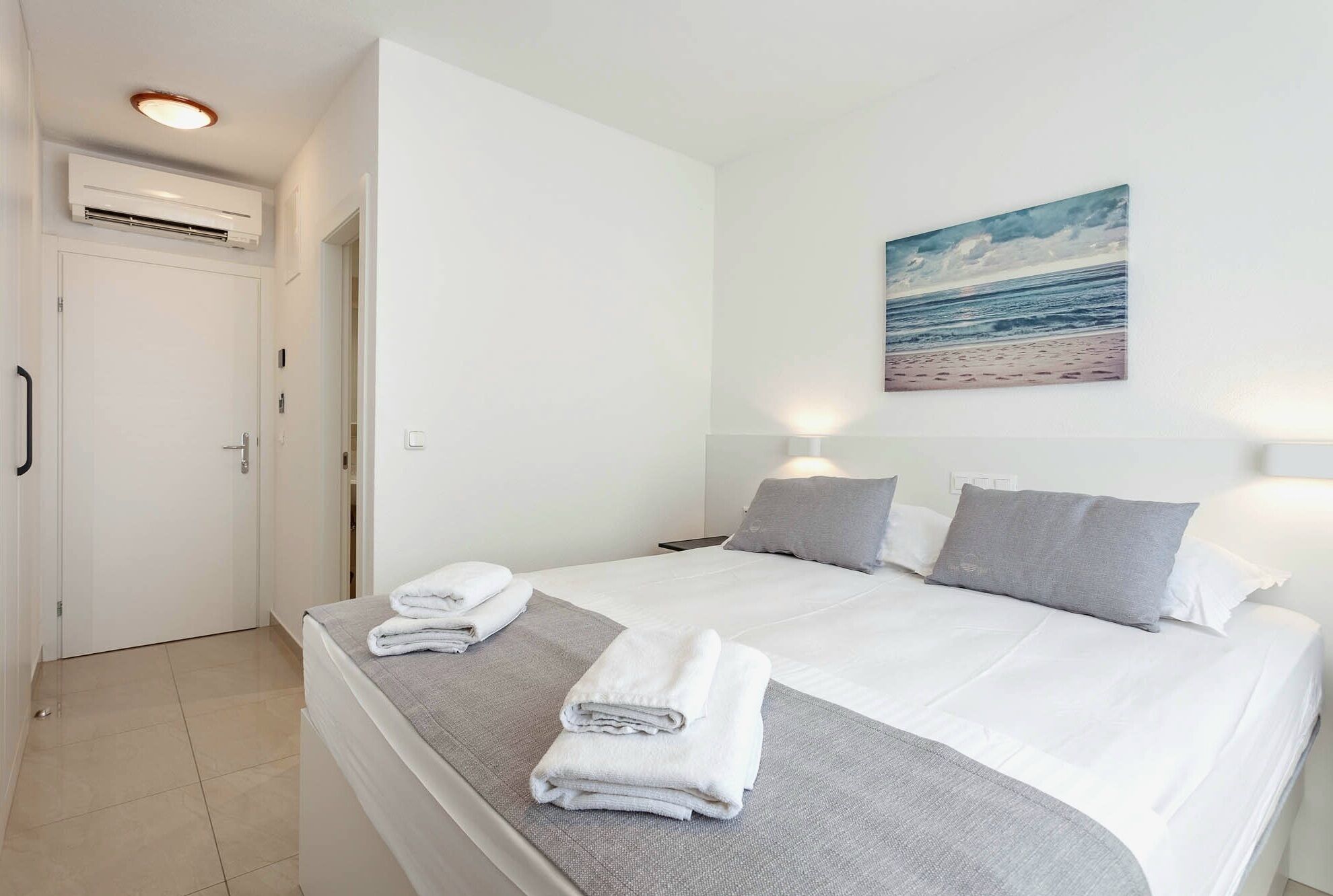 A hotel room with a bed, two bedside lamps, a painting of a beach, and a white door.
