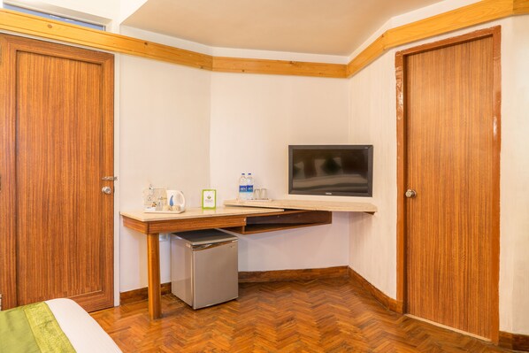 A small room with wooden flooring, a bed, a desk with a television, and a small refrigerator.