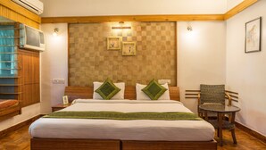 Una habitación de hotel con una cama de madera, dos almohadas verdes, una mesita redonda y una silla de mimbre.