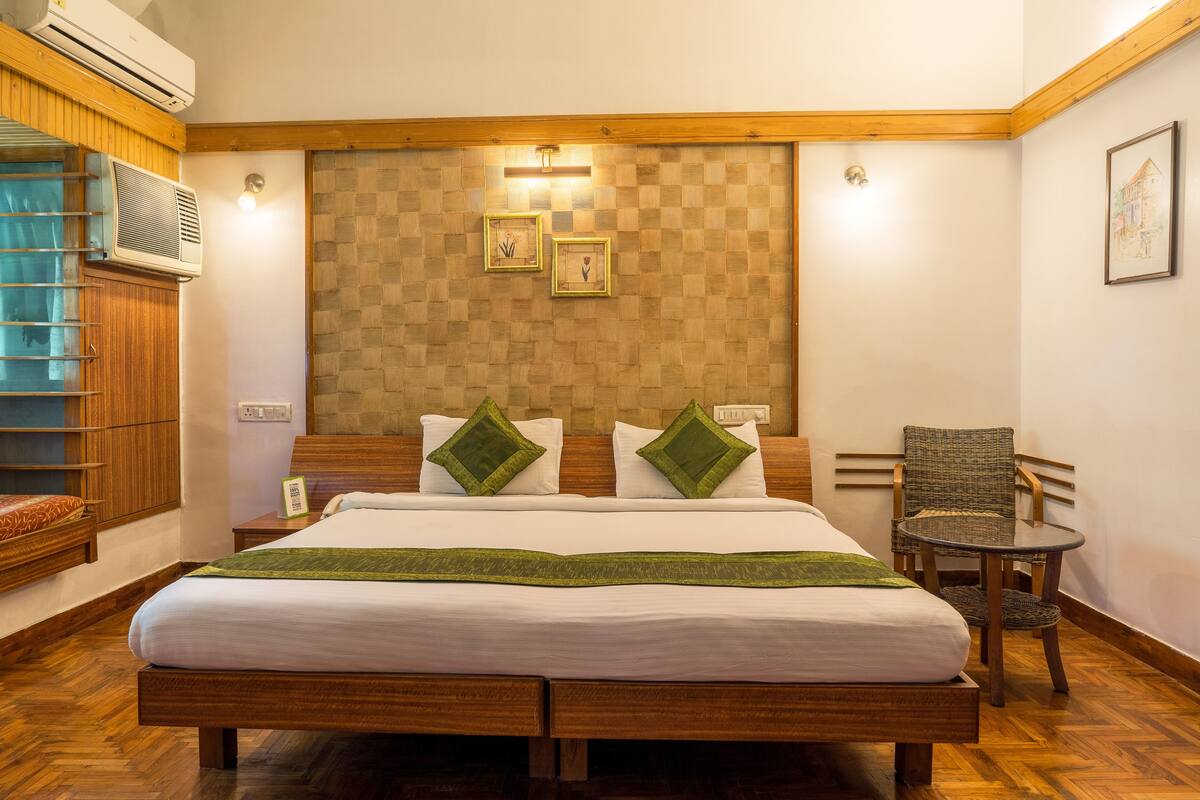 A hotel room with a wooden bed, two green pillows, a small round table, and a wicker chair.