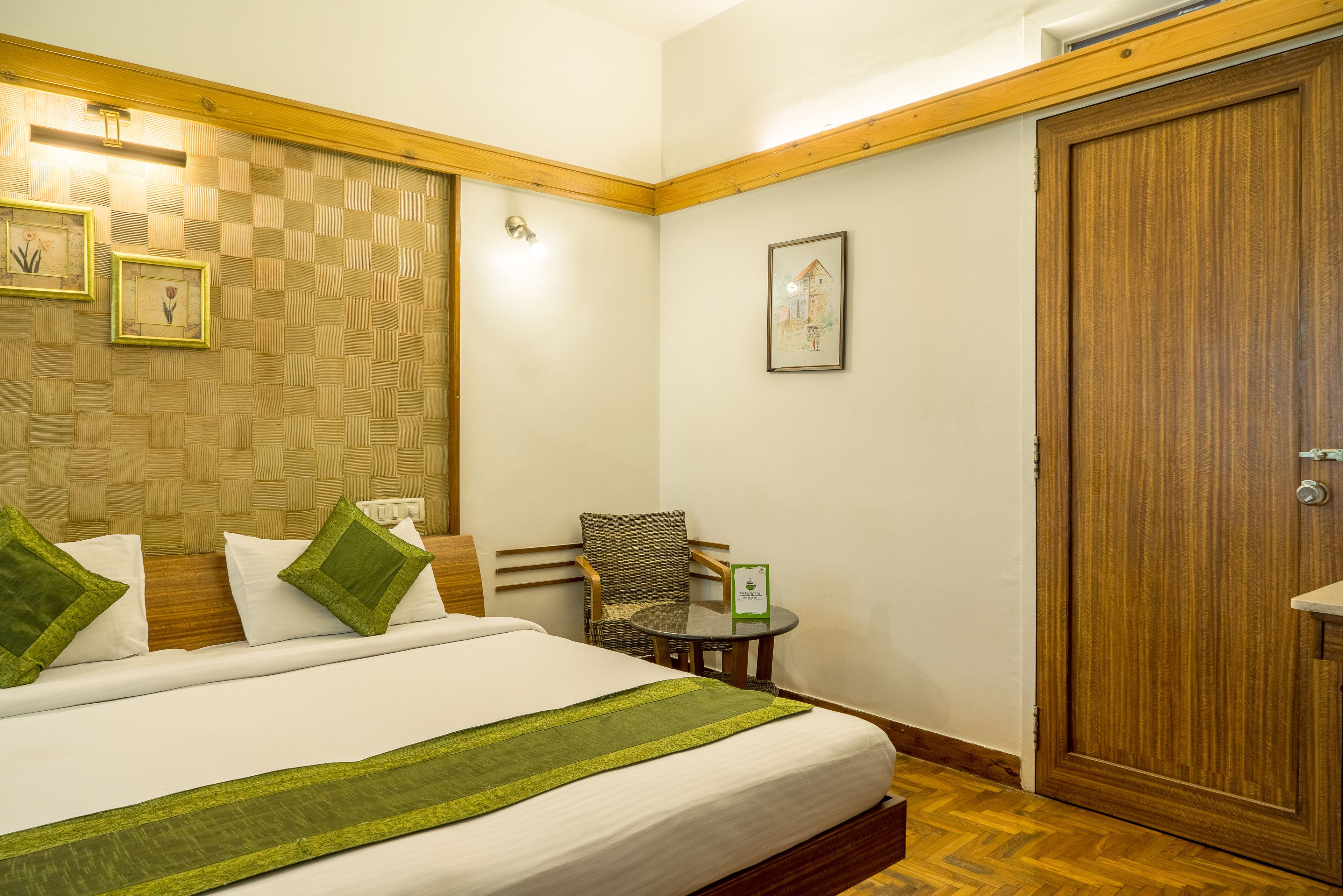 Quarto de hotel com uma cama grande, uma mesinha redonda, uma cadeira e uma porta de madeira.