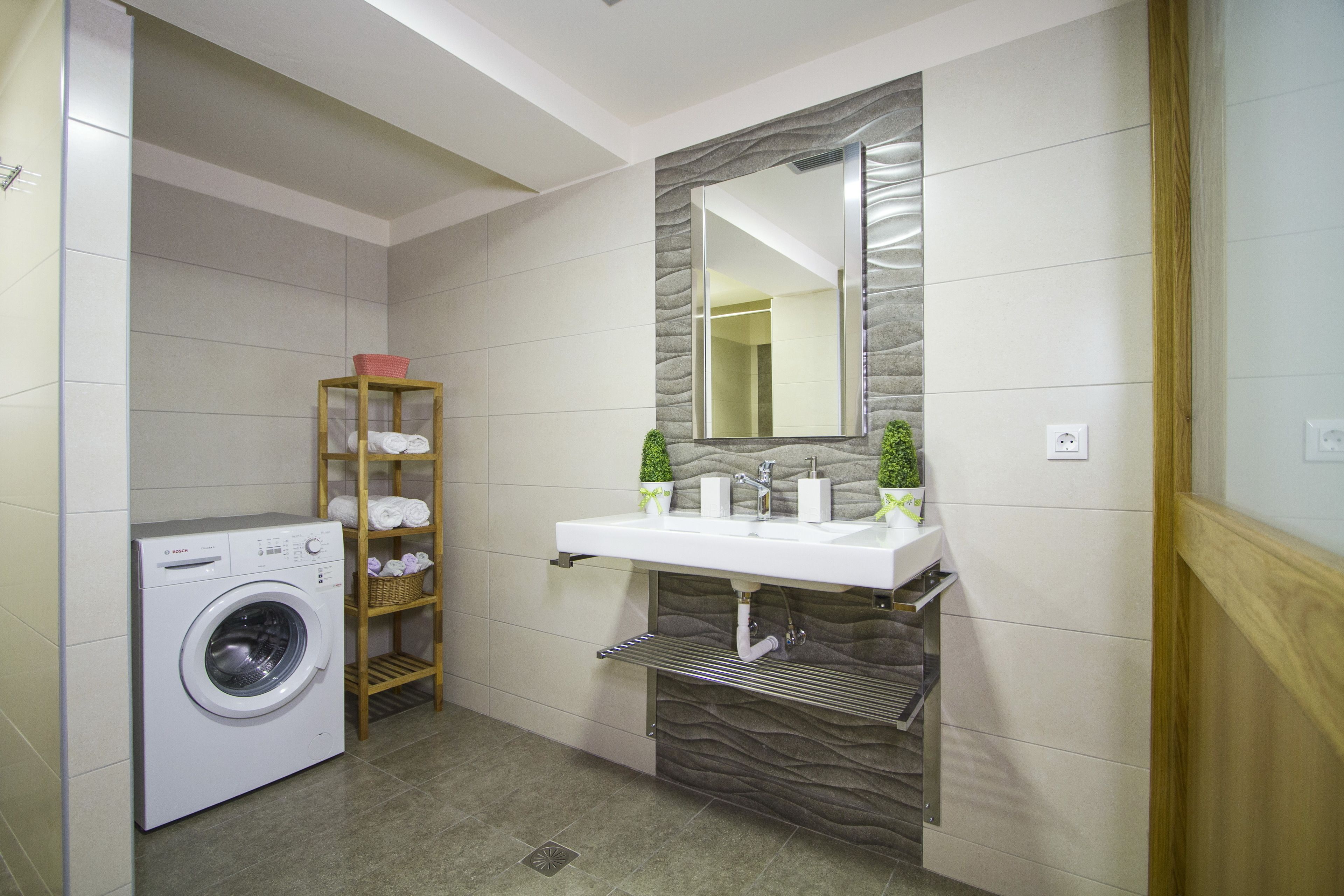 A modern bathroom with a washing machine, a wooden shelf with towels, a sink with a mirror, and a stone accent wall.