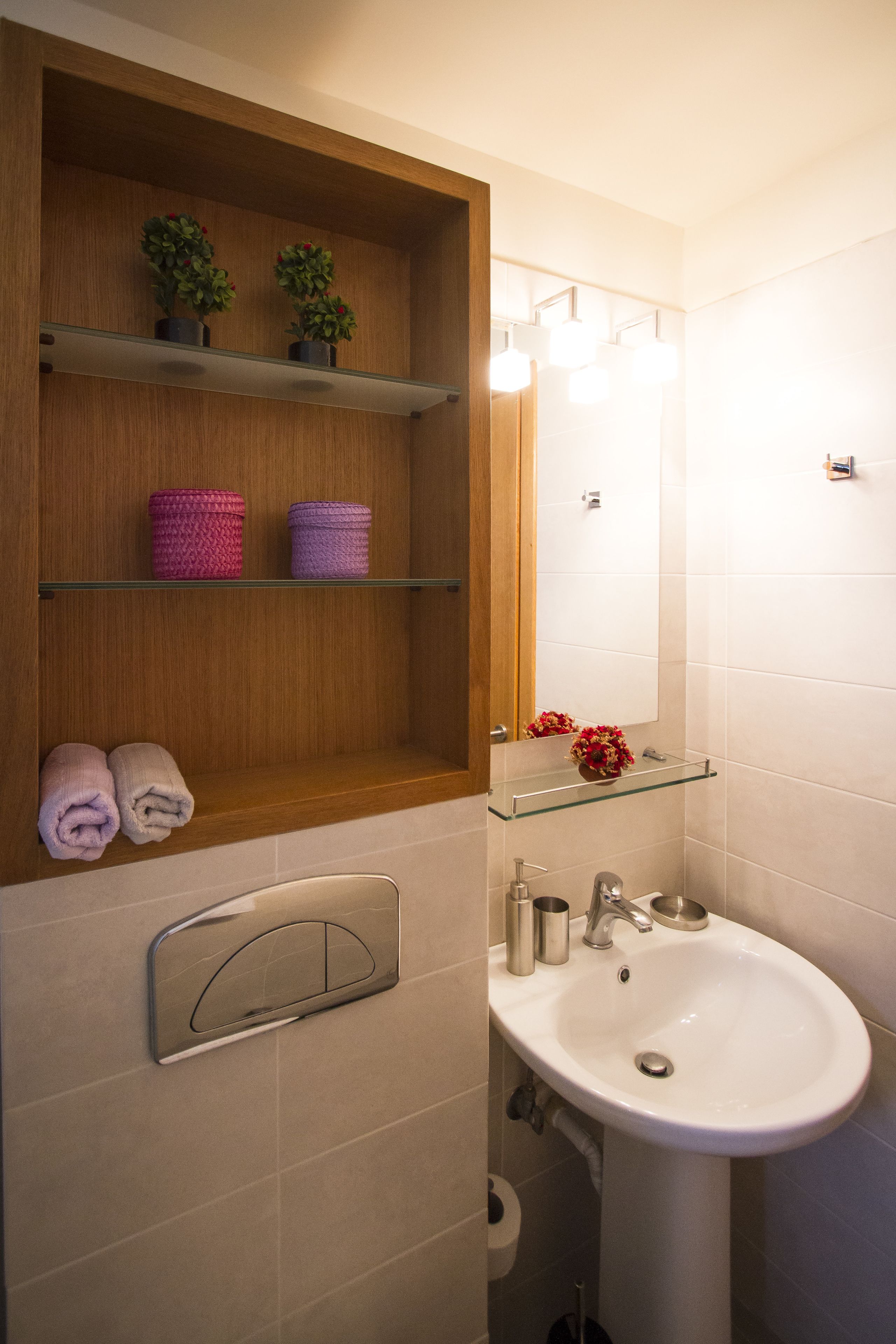 A bathroom with a wooden shelf holding towels and potted plants, a glass shelf with a vase, a white sink, and a mirror with lights.