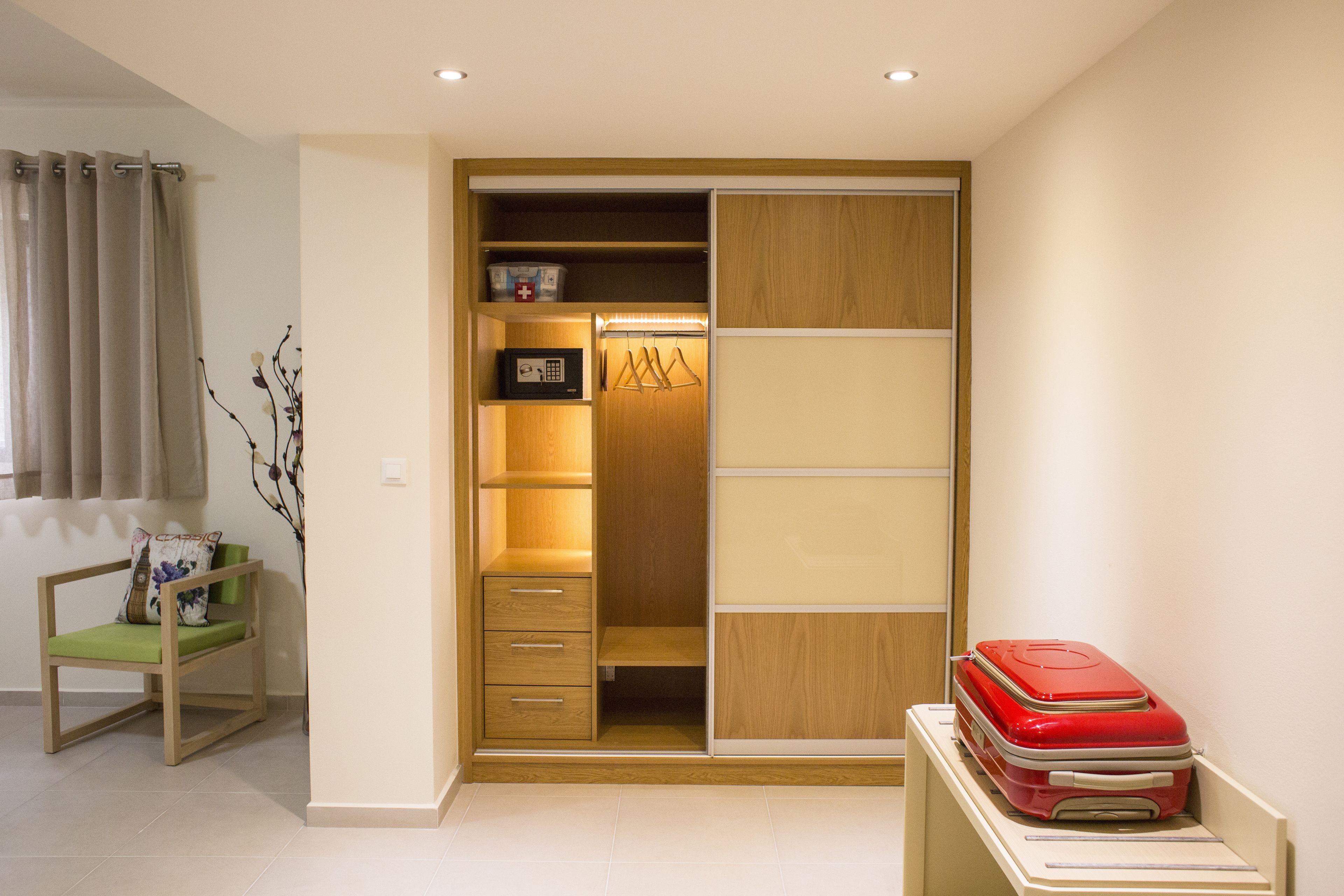 A room with a wooden wardrobe, a green chair, and a red suitcase on a table.