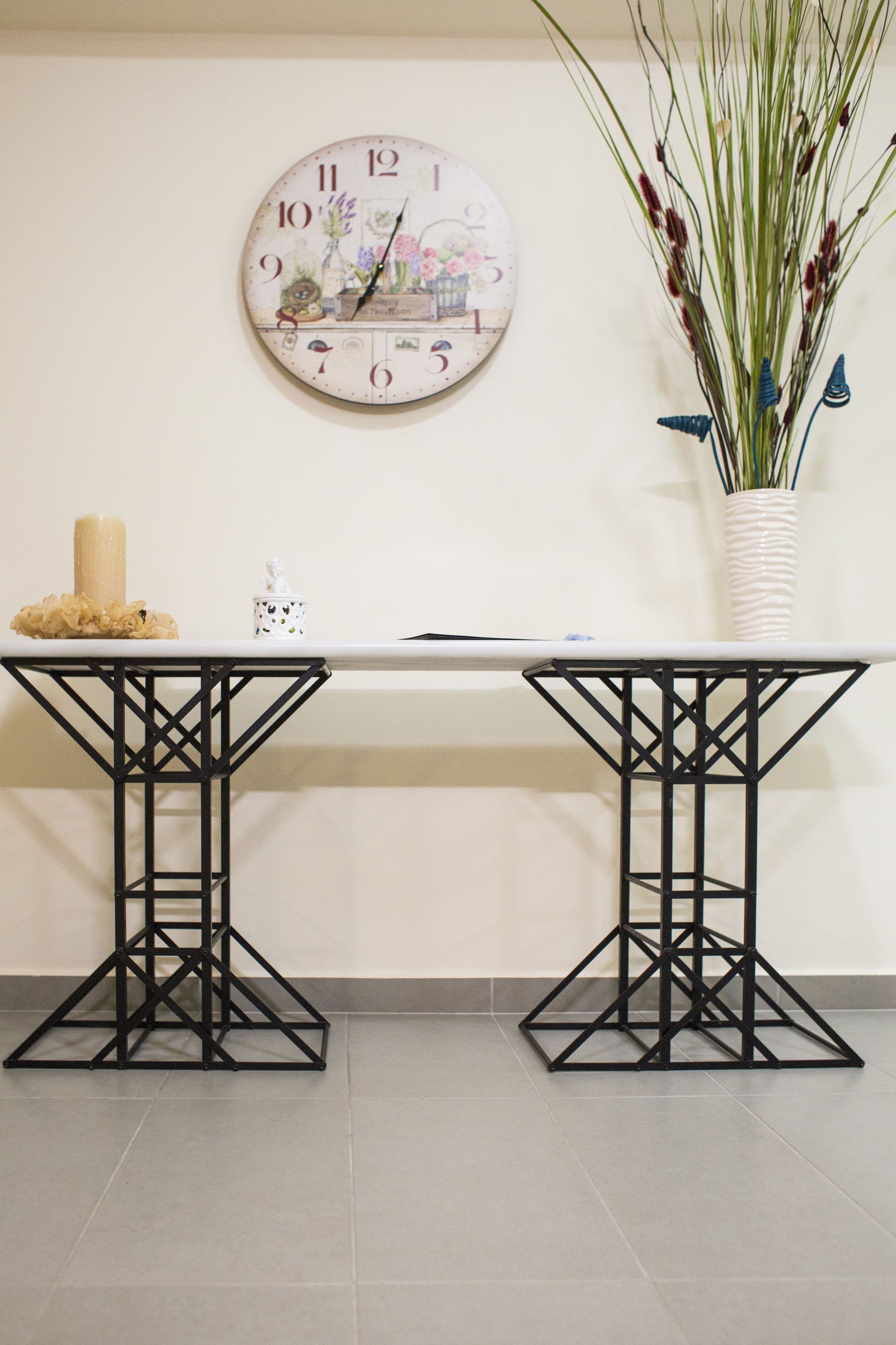 A white table with a black metal frame, a decorative clock with floral designs, and a vase with tall plants on the wall.