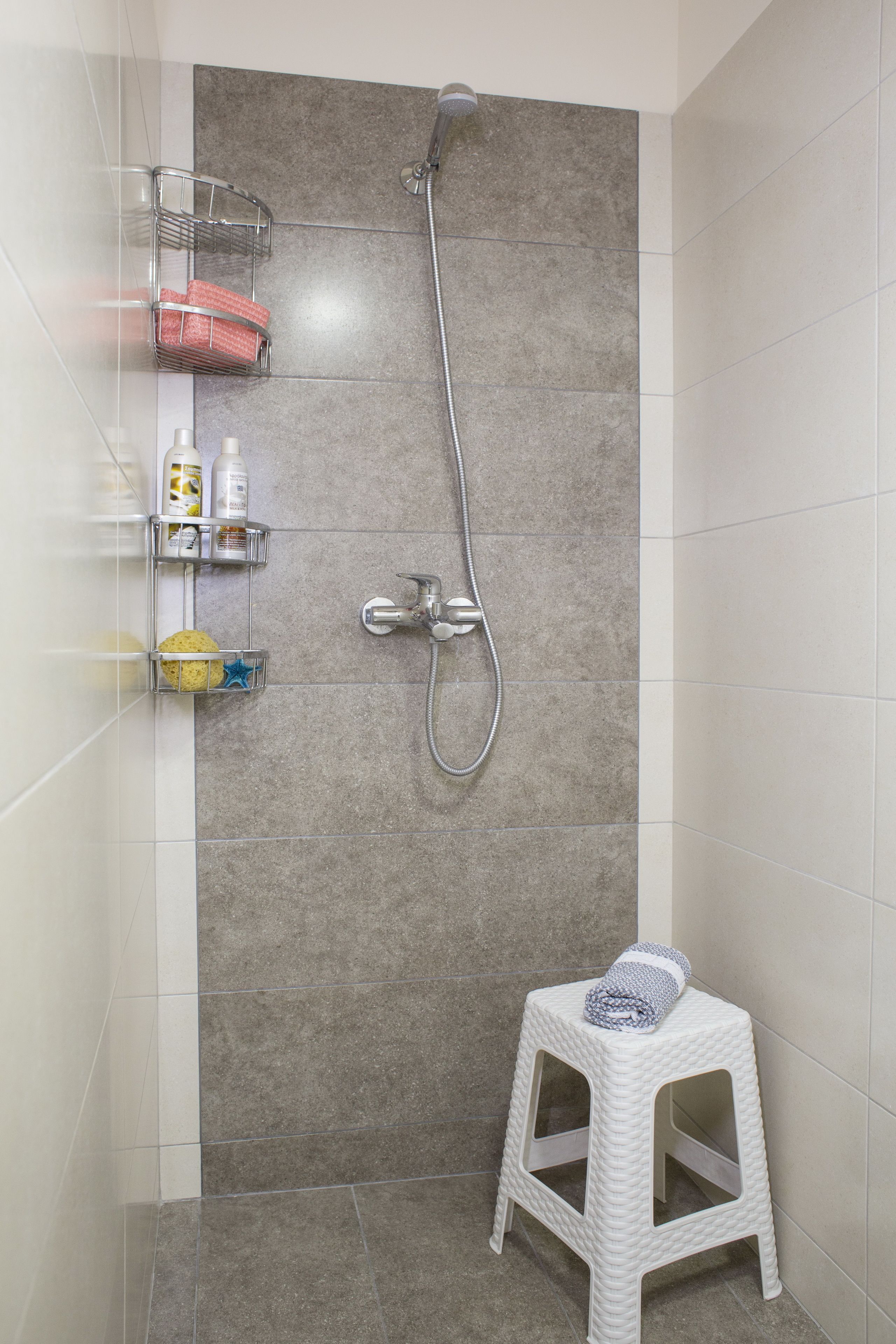 Zona de ducha con cabezal de ducha de mano, un taburete pequeño y repisas con artículos de aseo.