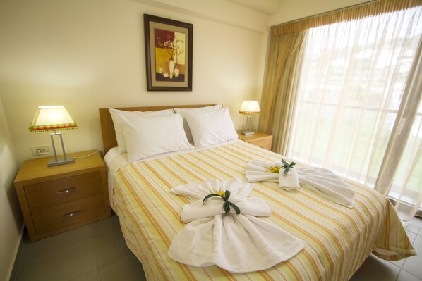 A neatly made bed with striped bedding, two bedside tables with lamps, a framed picture on the wall, and a large window with curtains.