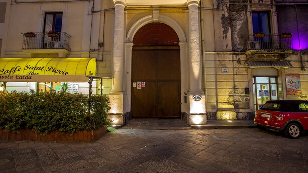 A building entrance with a wooden door, a yellow awning, and a red car parked nearby. - Sicilia Etna Mare (Riposto)