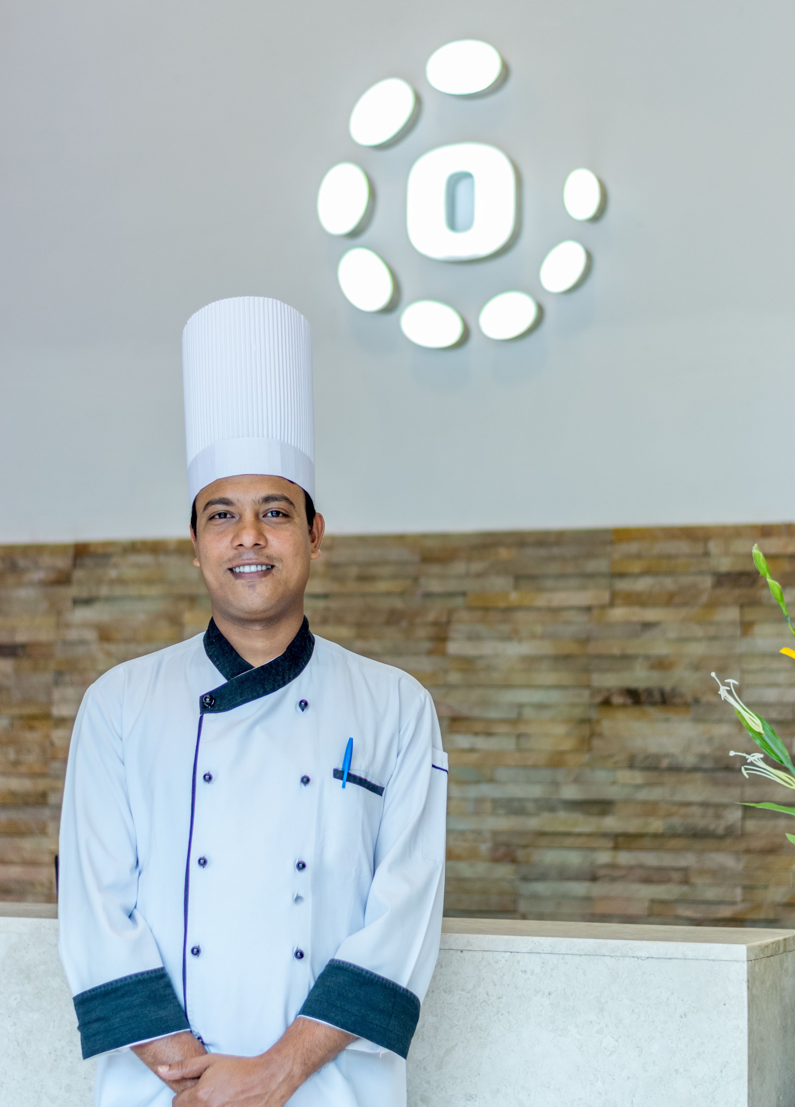 A person in a chef's uniform standing in front of a reception area with a modern light fixture and a stone wall.