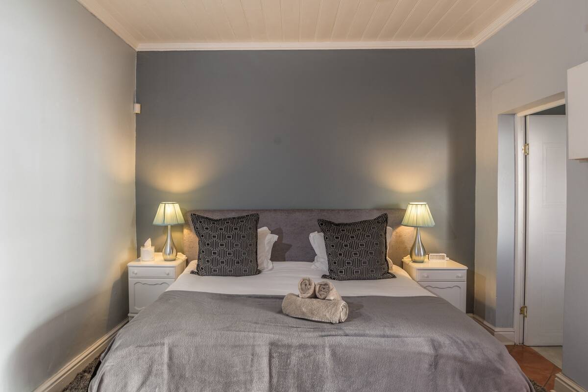 A neatly made bed with a grey blanket and two pillows, two bedside lamps, and a small statue on the bed.