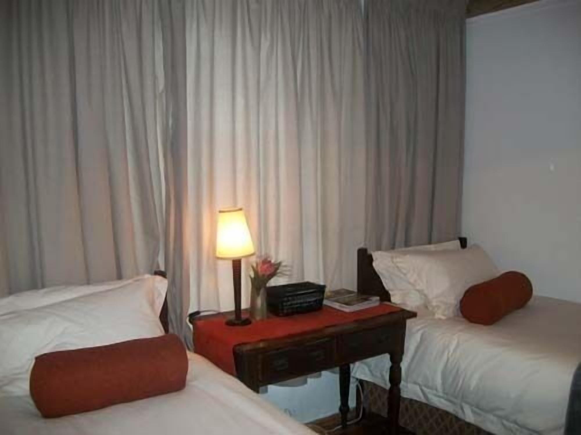Habitación de hotel con dos camas, una mesita de noche con lámpara, y una pequeña mesa con un libro y un florero.