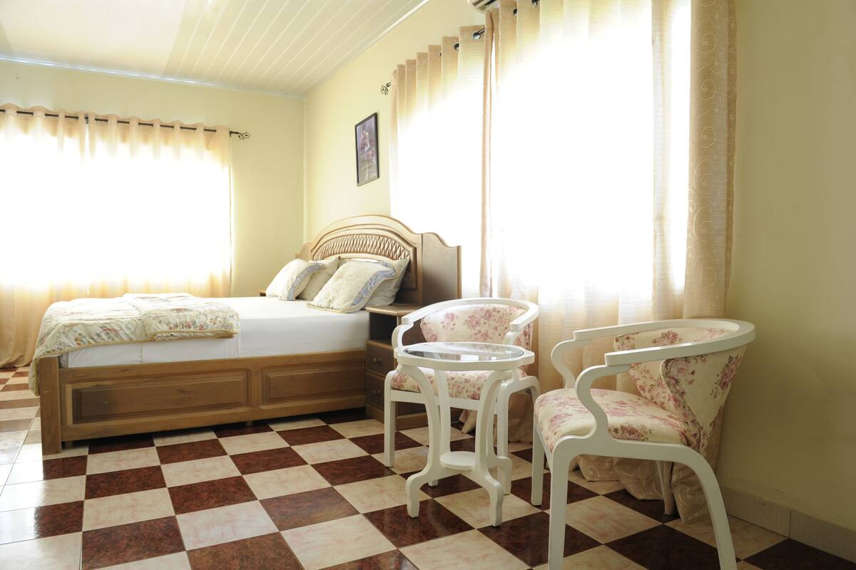 Une chambre avec un sol à carreaux, un lit doté d’une tête de lit en bois, deux chaises blanches à motifs floraux et une petite table ronde.