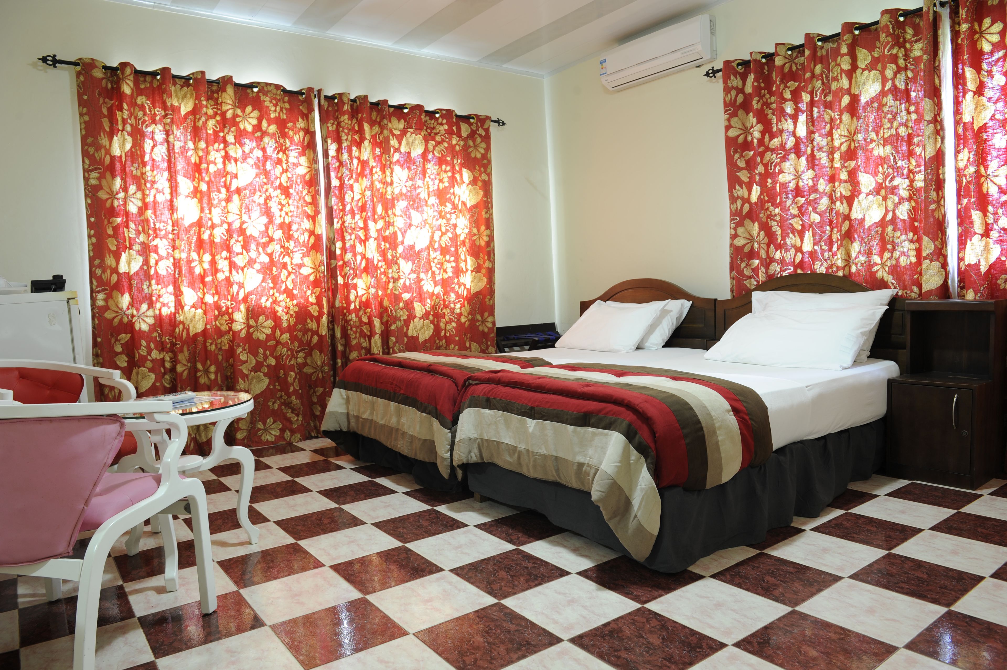 A hotel room with a checkered floor, a bed with a red and beige bedspread, a wooden headboard, a bedside table, a nightstand, and a chair.