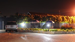 A paved parking area with a white SUV parked, surrounded by a landscaped garden with trees and shrubs, and outdoor seating under umbrellas. - Mount Pleasant Inns & Apartment (Obosomase)