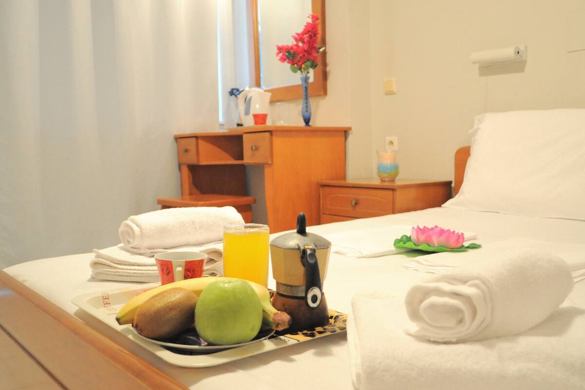 A neatly made bed with a rolled white towel, a plate with fruits and a mug, a bedside table with a lamp, and a nightstand with a vase of flowers.