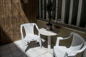 Two white plastic chairs and a small round table with a potted plant on a tiled patio. - Greta Apartments (Hersonissos)