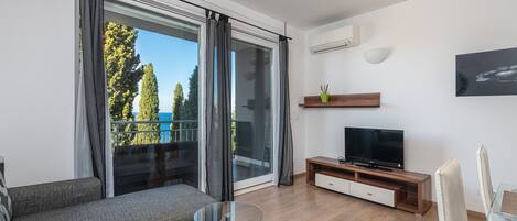 Ein modernes Wohnzimmer mit einer Couch, einem Glastisch, einem Fernseher und einem Balkon mit Vorhängen.