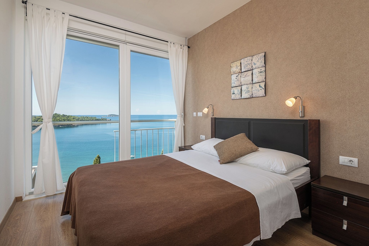 A bedroom with a large bed, a nightstand, a lamp, and a view of the sea.
