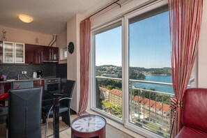 A modern kitchen with a dining area, large windows with a view of a lake and buildings, and a red leather chair. - Apartments Didan (Konavle)
