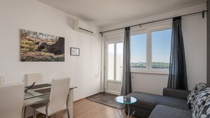 A modern living room with a glass dining table, grey sofa, and a large window with curtains. - Apartments Didan (Konavle)