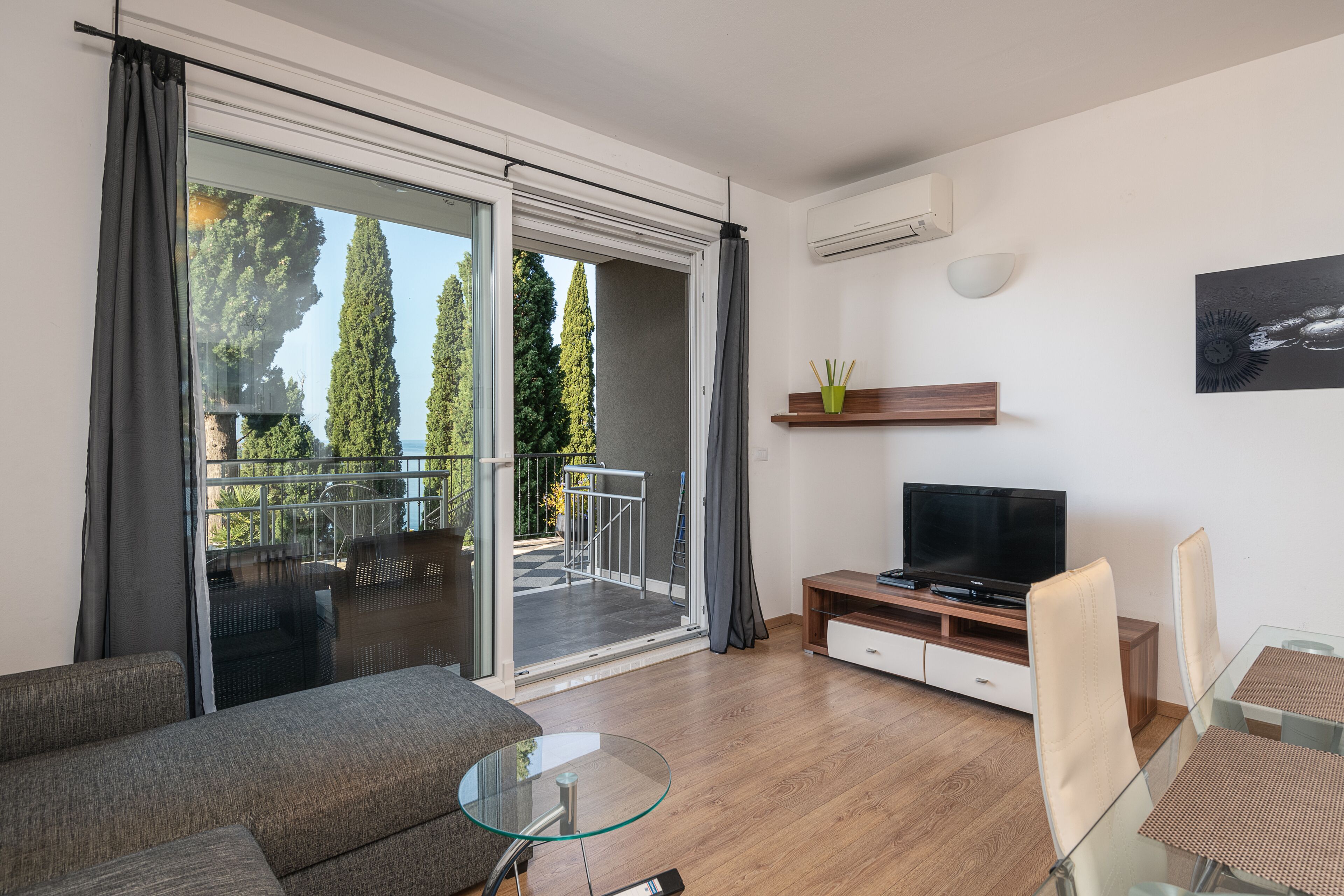 Un salón moderno con sofá, mesa de centro de cristal, televisor y balcón con vistas a árboles.