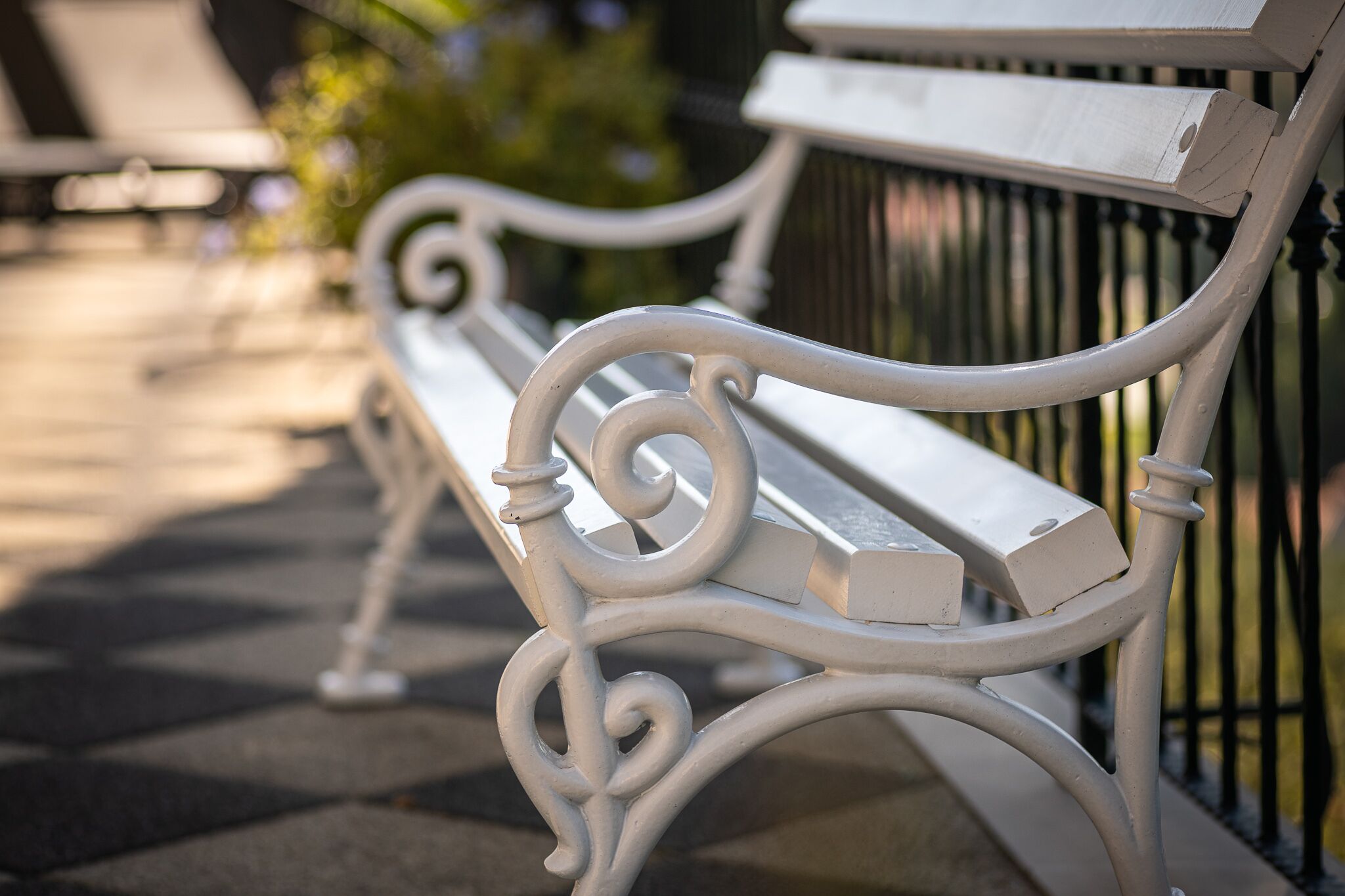 A white metal bench with ornate scrollwork, positioned next to a metal fence.
