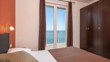 A bedroom with a bed, a nightstand, a lamp, a wardrobe, and a sliding glass door leading to a balcony with a view of the sea.