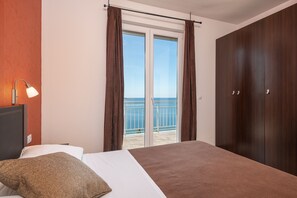 A bedroom with a bed, a nightstand, a lamp, a wardrobe, and a sliding glass door leading to a balcony with a view of the sea. - Apartments Didan (Konavle)