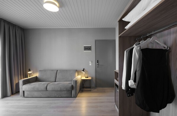 A modern hotel room with a grey sofa, bedside tables with lamps, a wardrobe with clothes, and a door. - Skianthion (Skiathos)